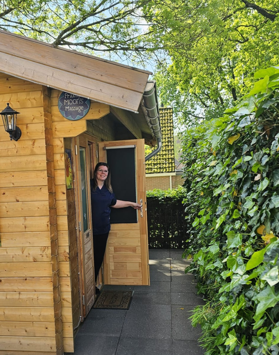 Buitenkant van de houten praktijkcabine Moon's Massage in Apeldoorn, met naam op het bord boven de deur en uitnodigende deuropening