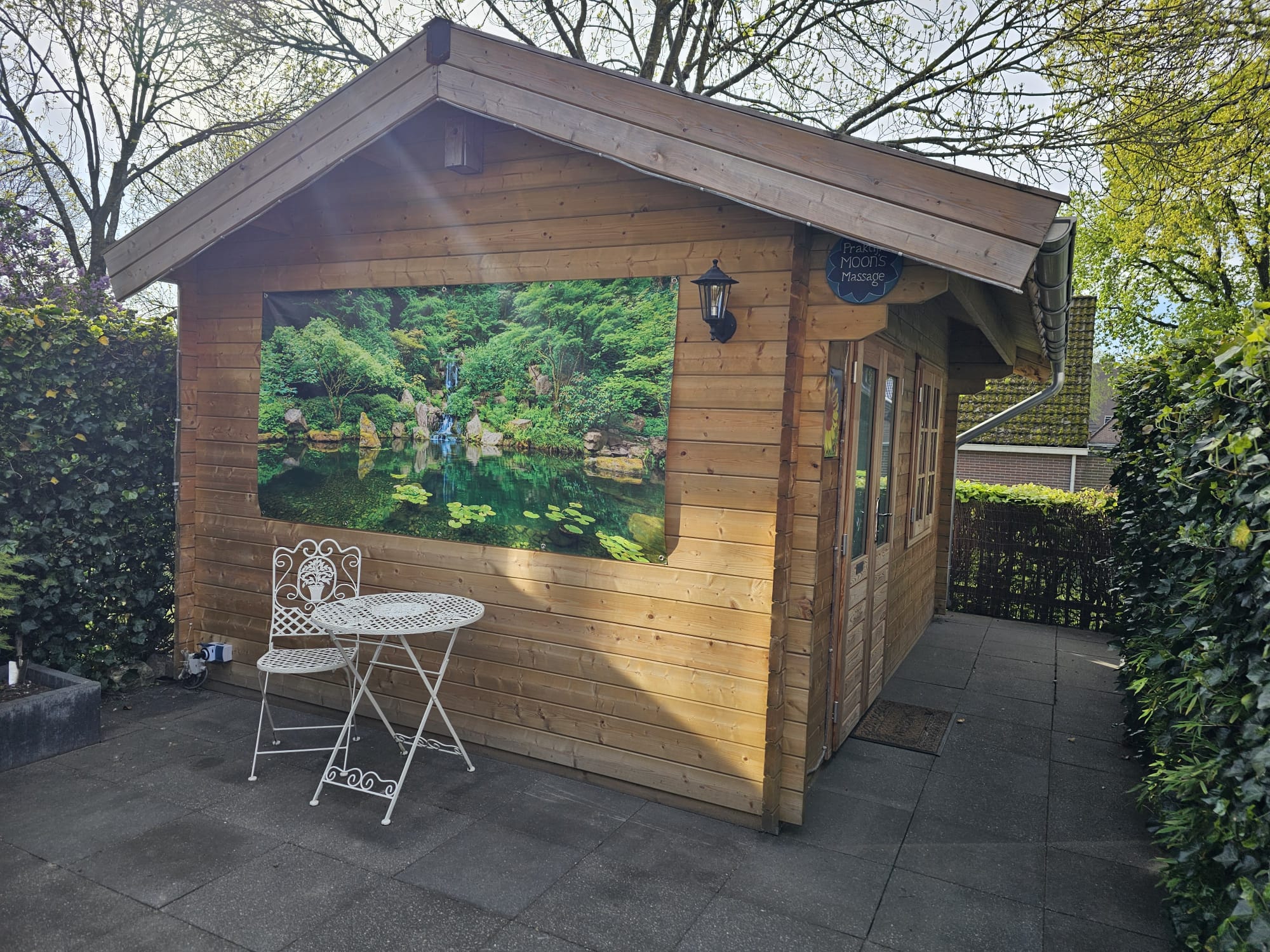 Buitenaanzicht van Praktijk Moon's Massage: een knusse houten praktijkcabine met natuurbanner, bord en een wit zitje op het stenen terras